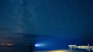 Cilvēks ar lukturīti stāv uz jūras krasta tumšā naktī zem zvaigžņotām debesīm Latvijā.