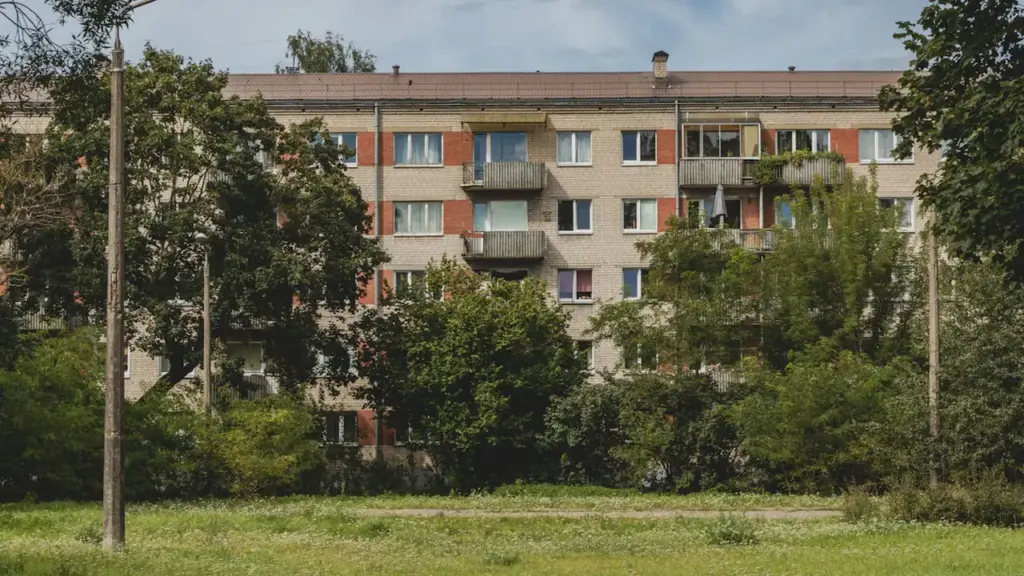 Tipiska daudzdzīvokļu māja Rīgas mikrorajonā, ko ieskauj koki un zaļa zāle.