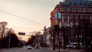 Automašīnas brauc pa Rīgas ielu pēcpusdienas gaismā ar pilsētas arhitektūru fonā.