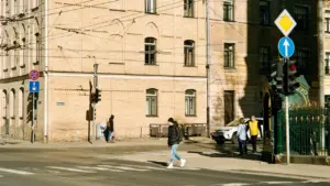 Cilvēki šķērso ielu Rīgā pie krustojuma ar satiksmes gaismām un ceļa zīmēm.