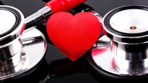 Medicīnas stetoskops un sarkana sirds uz galda, simbolizējot sirds veselības pārbaudes nepieciešamību.