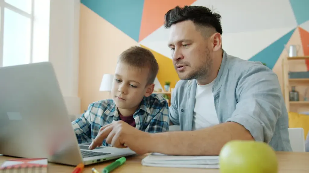Tēvs palīdz dēlam pildīt mājasdarbus pie datora mājās, smaidot un mācoties kopā.