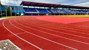 Sarkans vieglatlētikas skrejceļš ar baltām līnijām un skatītāju tribīnēm ar zilām sēdvietām stadionā.